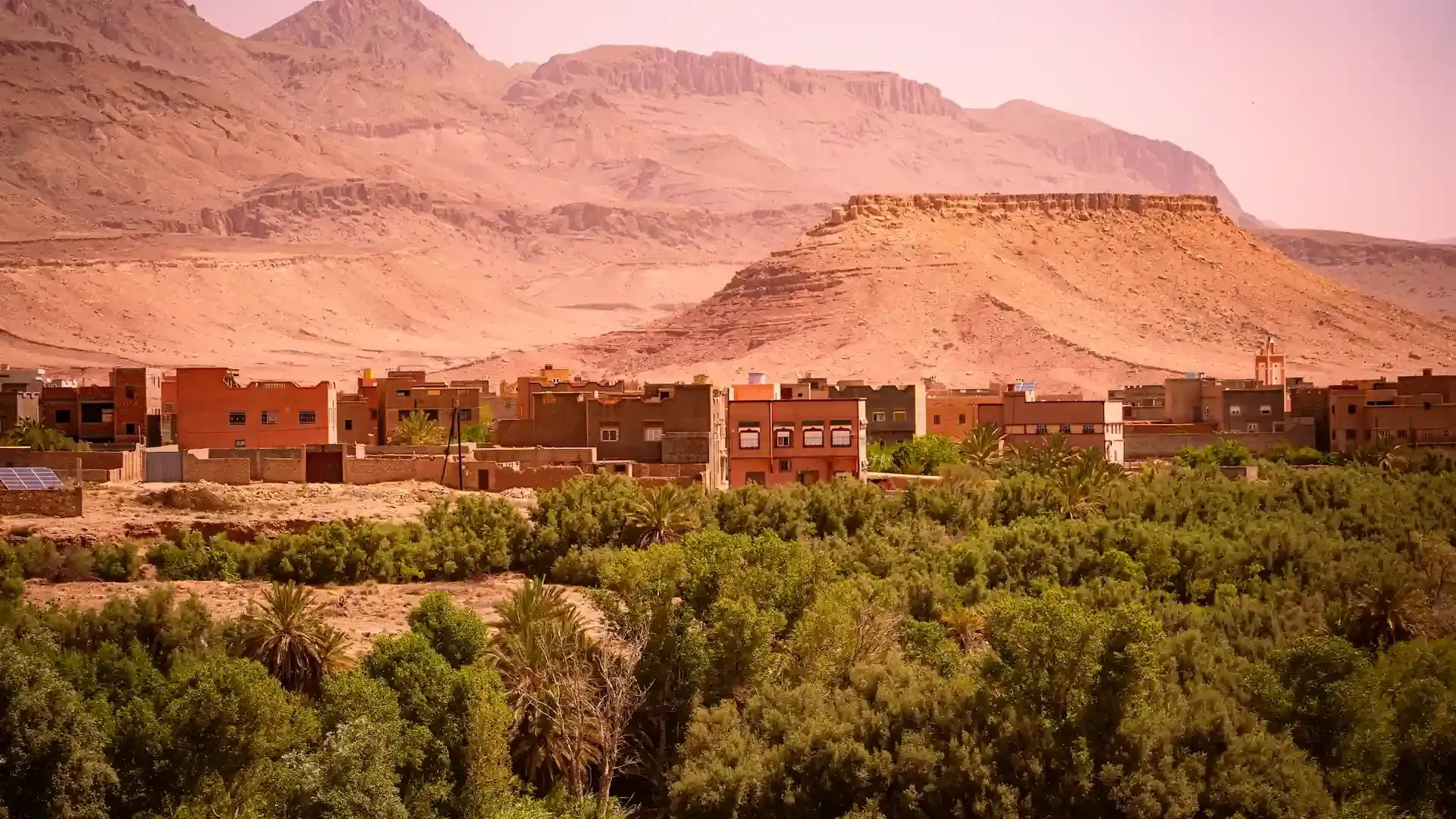Pueblo situado al pie de montañas áridas y color canela, con edificios en tonos tierra, sobre un exuberante follaje verde.