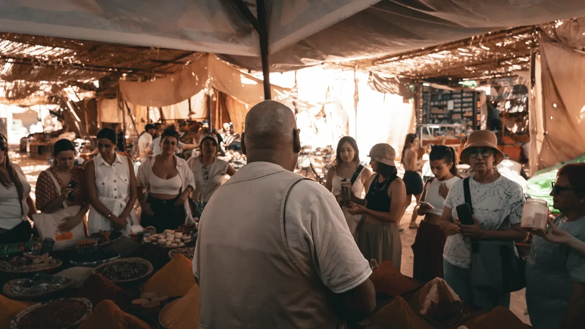 Un hombre habla con un grupo en un puesto de mercado al aire libre, con comida a la vista.