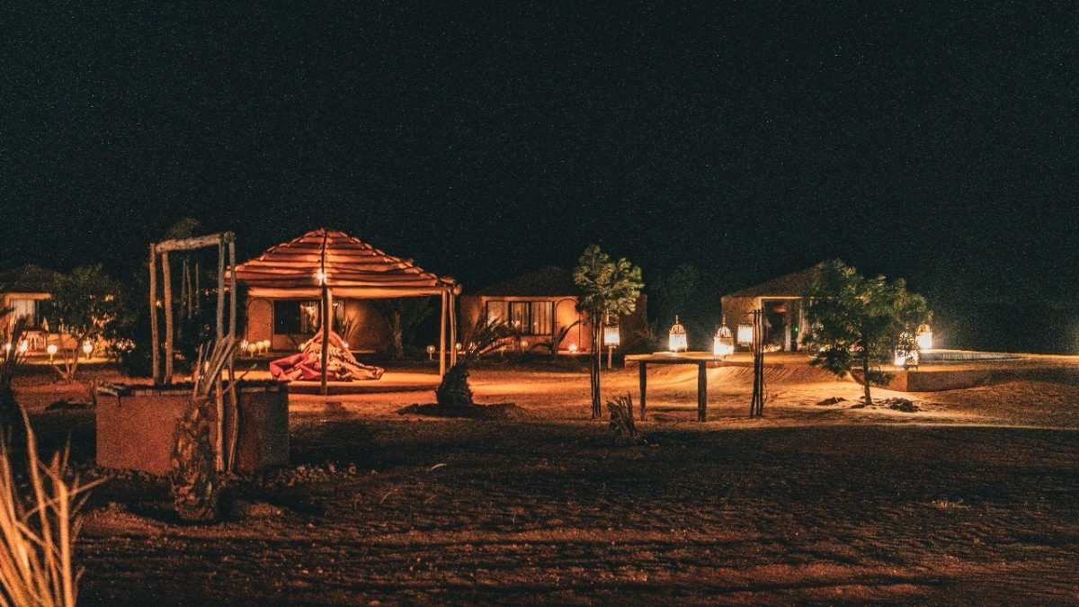 Cabañas iluminadas de noche, entorno desértico. Cielo oscuro, algunas estrellas visibles. Iluminación suave y cálida.