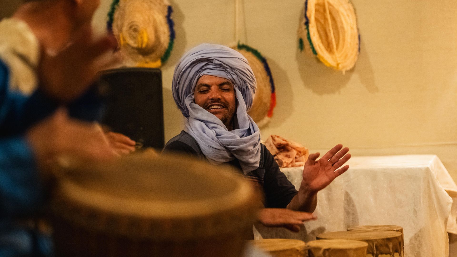 Un músico sonriente con un turbante azul claro toca tambores tradicionales en un espacio interior con sombreros de paja colgados en la pared.