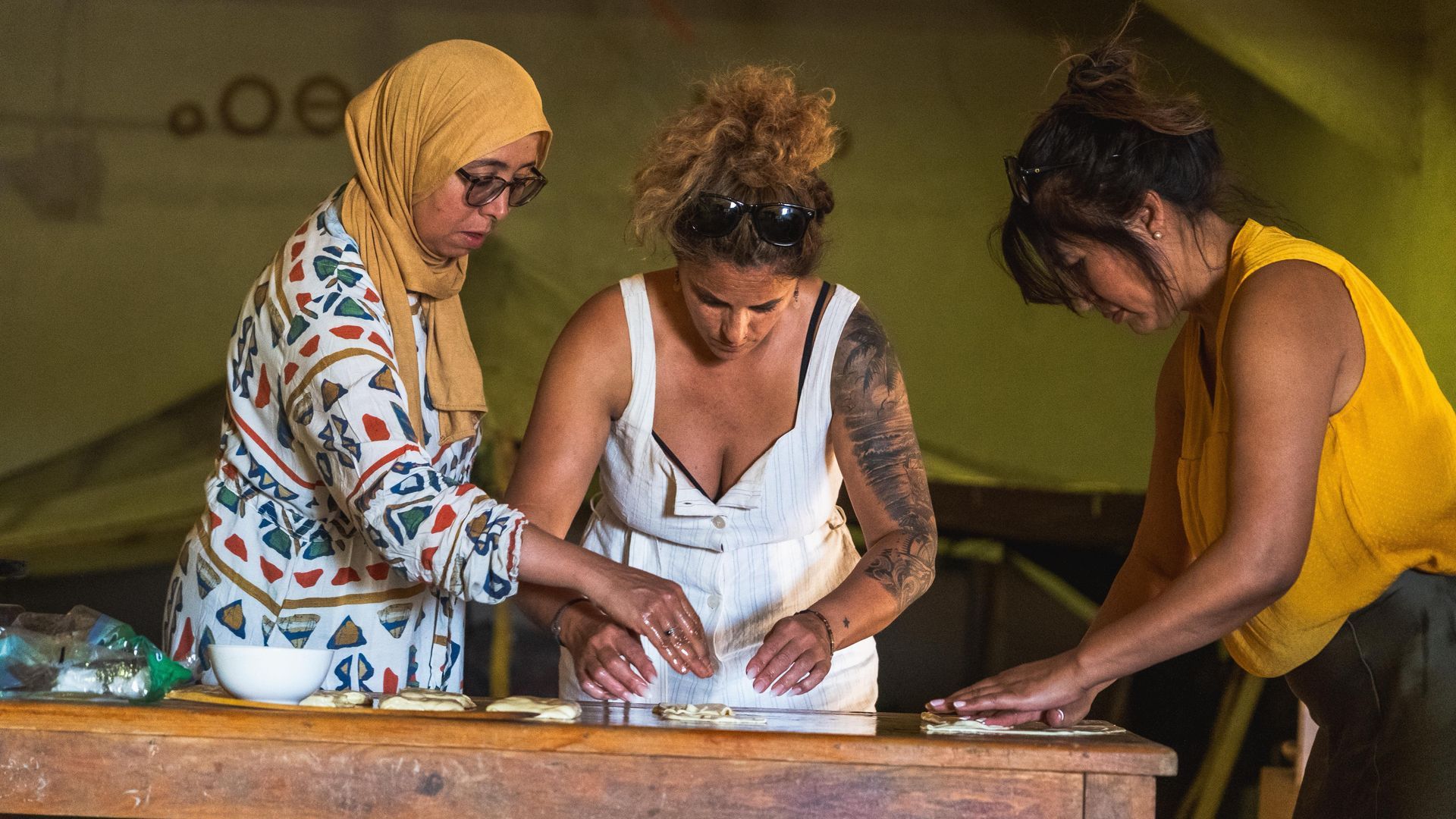 Tres personas están de pie junto a una mesa de madera, concentradas en dar forma a trozos de masa en un ambiente interior.