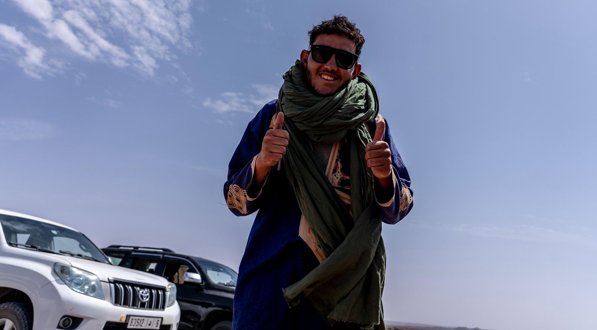 Una persona vestida con una túnica azul y una bufanda verde levanta ambos pulgares mientras usa gafas de sol al aire libre cerca de dos camionetas SUV.