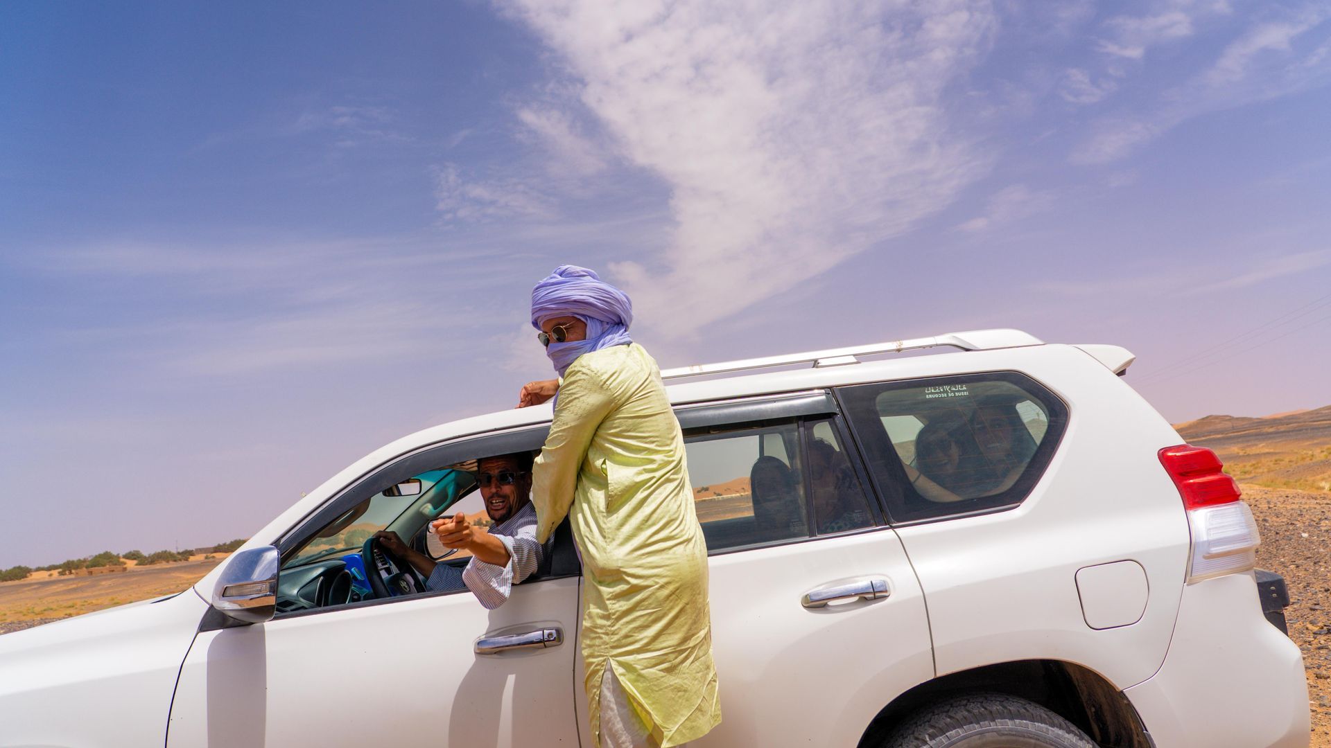 Una persona vestida con una túnica y un tocado de color amarillo claro está de pie junto a un SUV blanco, hablando con el conductor a través de la ventanilla.