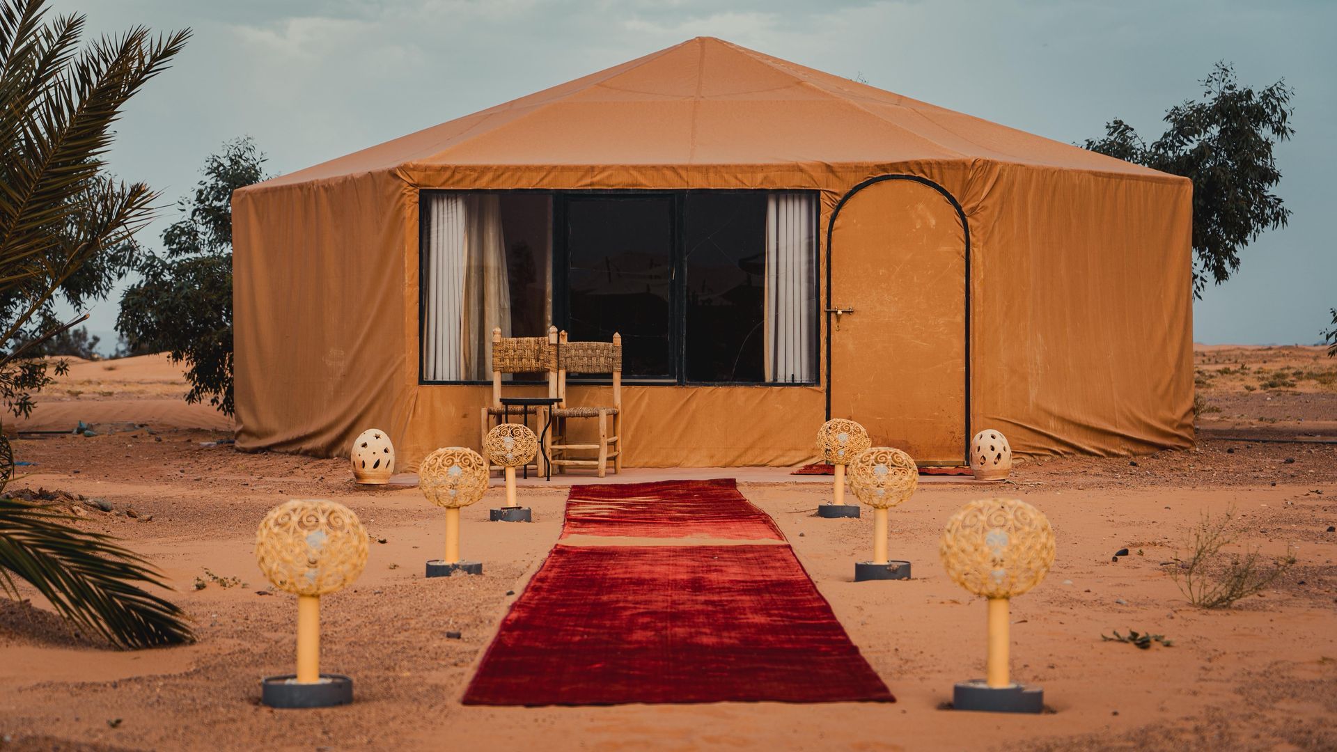 Una tienda de glamping en el desierto con una alfombra roja que conduce a la entrada, flanqueada por faroles de mimbre iluminados sobre suelo arenoso.