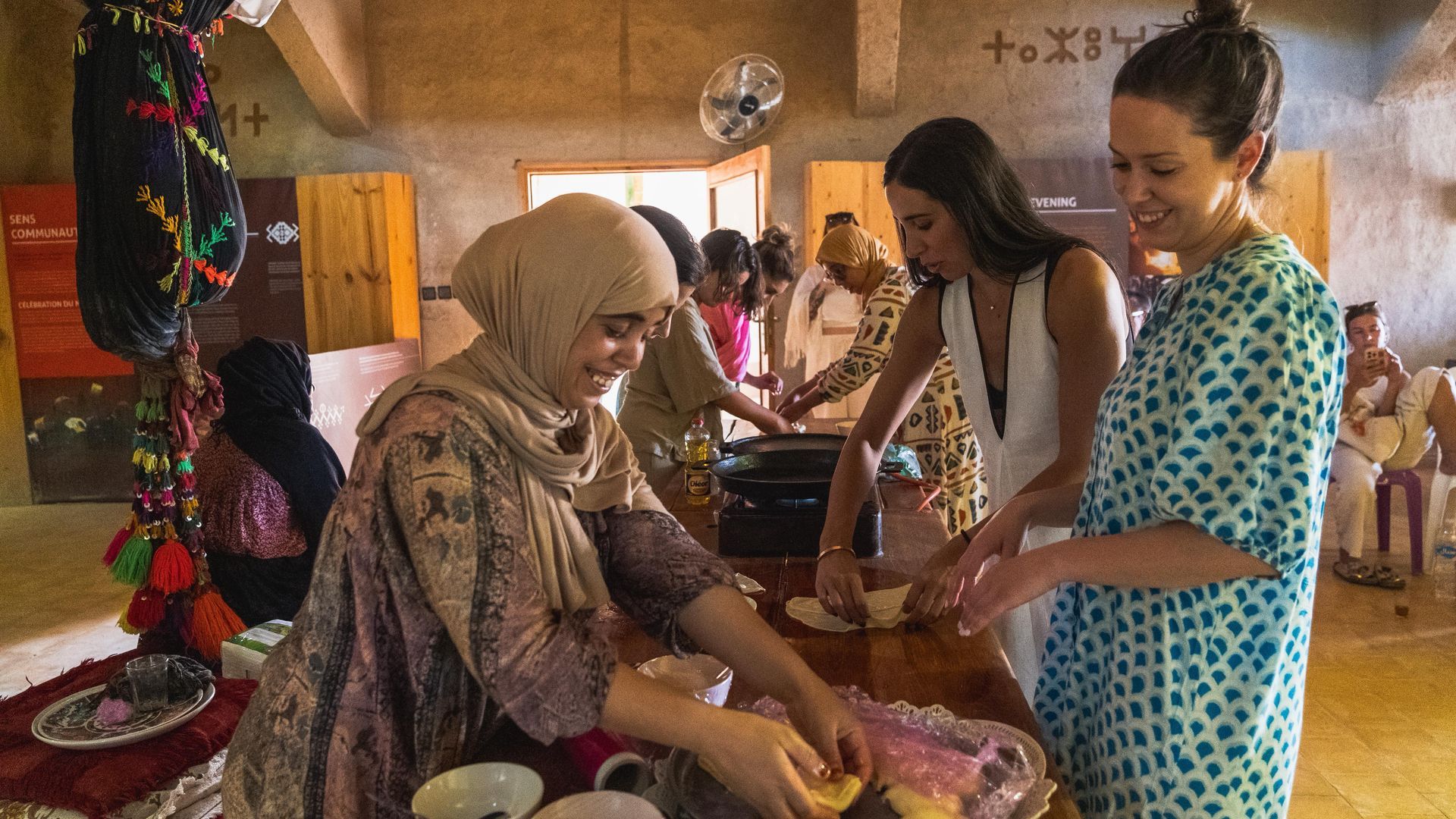 Varias personas trabajan juntas en una habitación soleada, preparando comida en una mesa de madera.