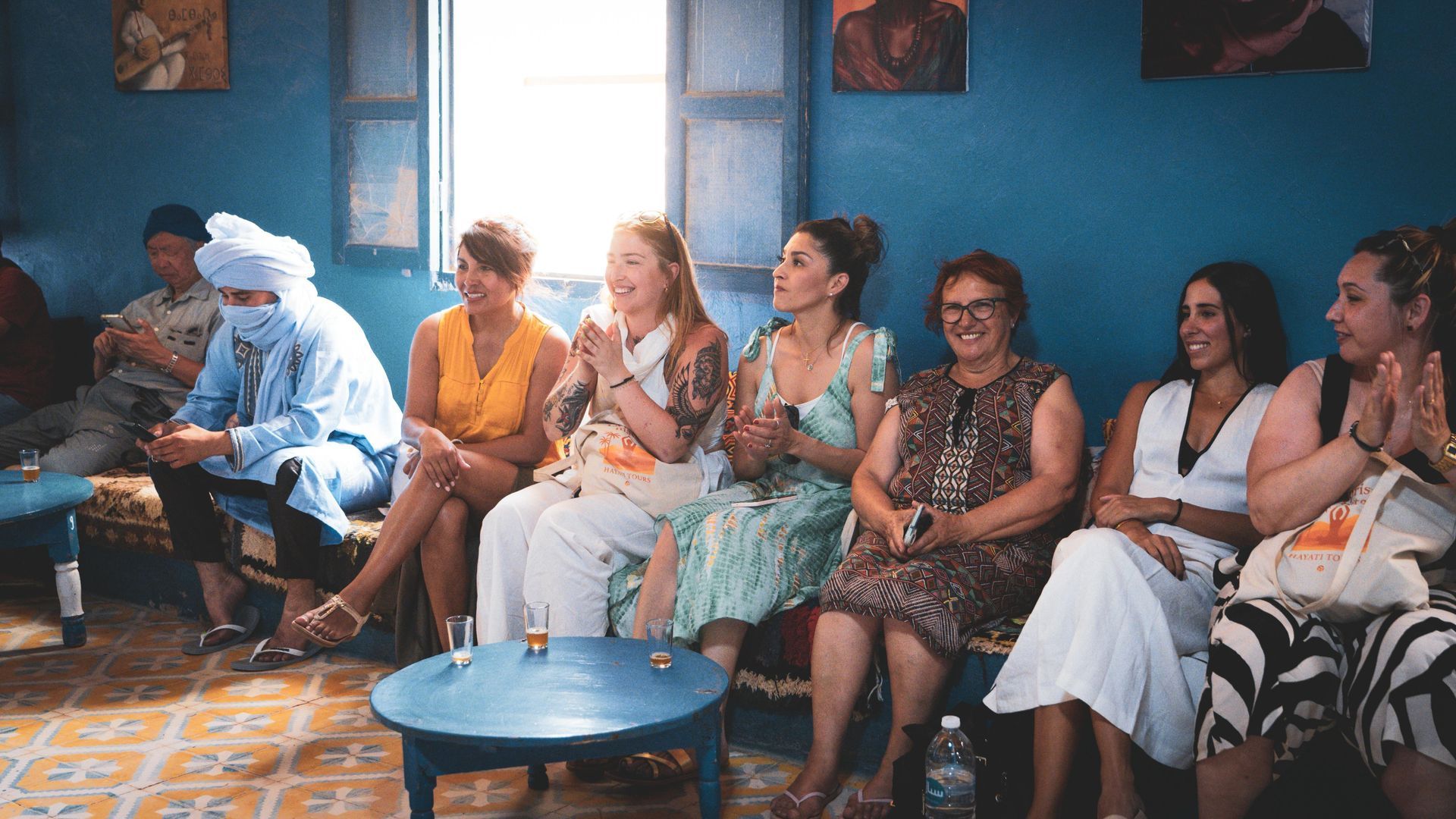 Un grupo diverso de personas sentadas en sillas en una sala de un azul vibrante, aplaudiendo y viendo una actuación.