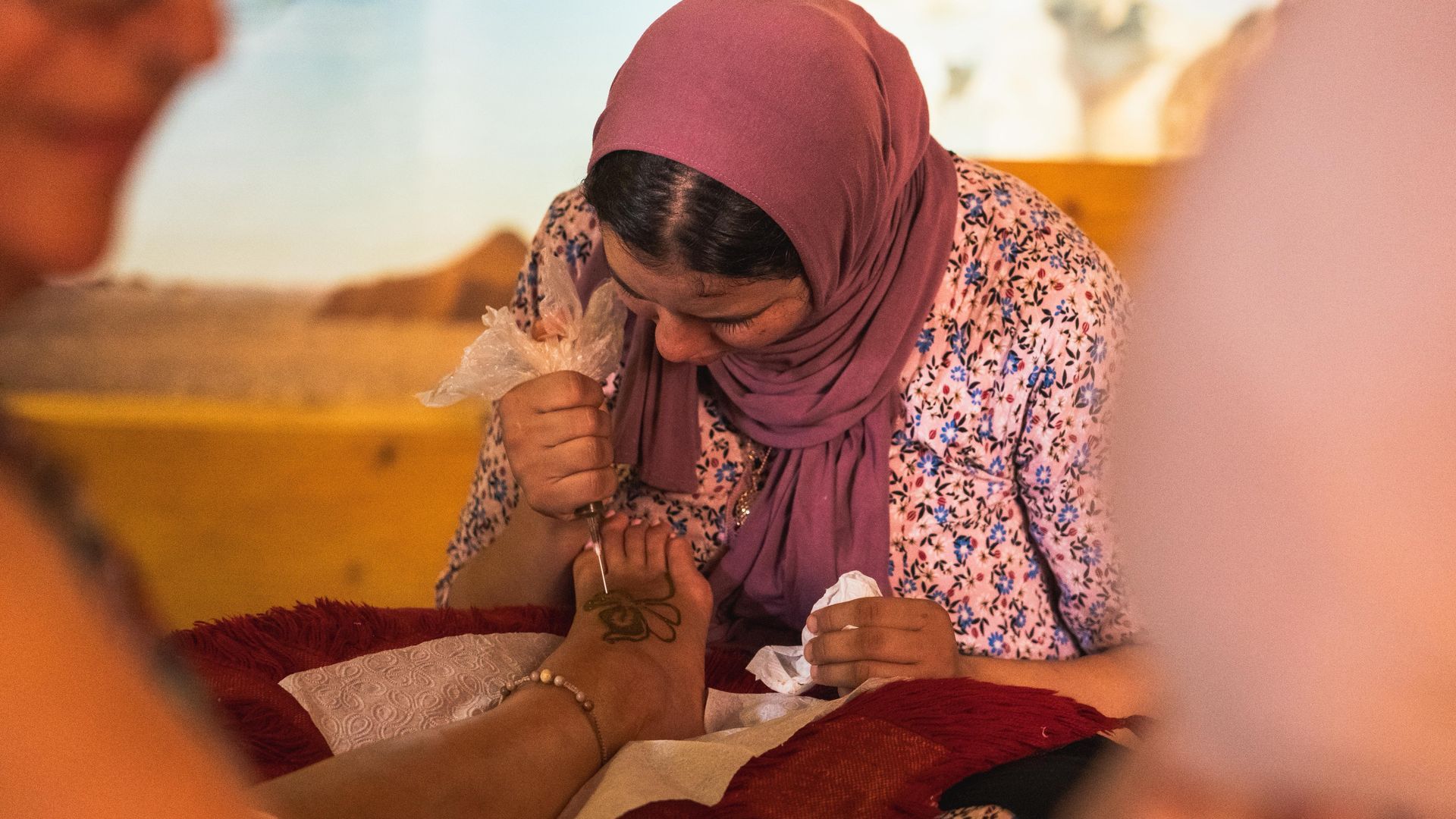 Una persona que lleva un pañuelo rosa en la cabeza aplica cuidadosamente henna en el pie de otra persona con un pequeño cono aplicador.
