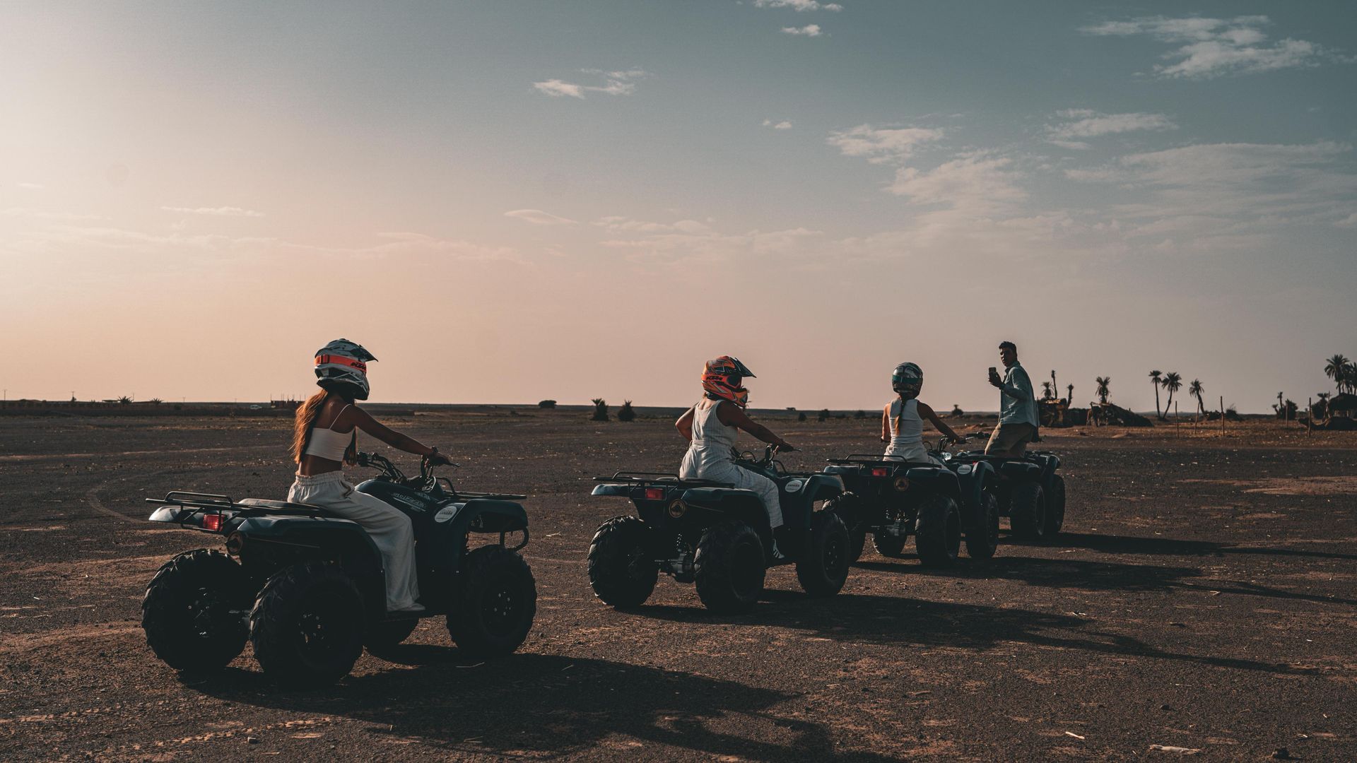 Cuatro personas montando vehículos todoterreno por un paisaje llano y polvoriento bajo un cielo despejado al atardecer.