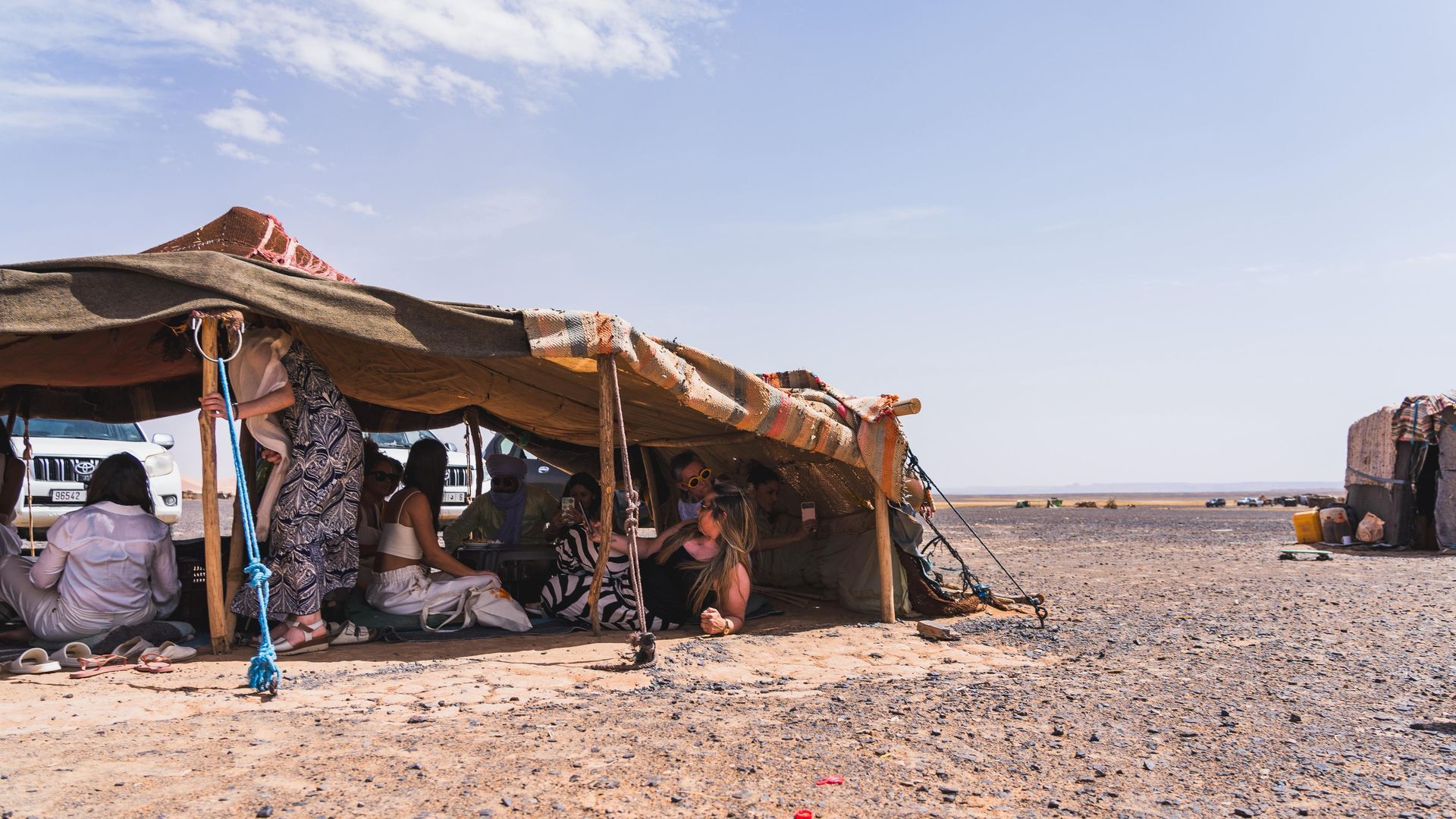 Un grupo de personas se sienta bajo una gran tienda de campaña tejida, de estructura baja, en un paisaje desértico llano y árido, bajo un cielo azul despejado.