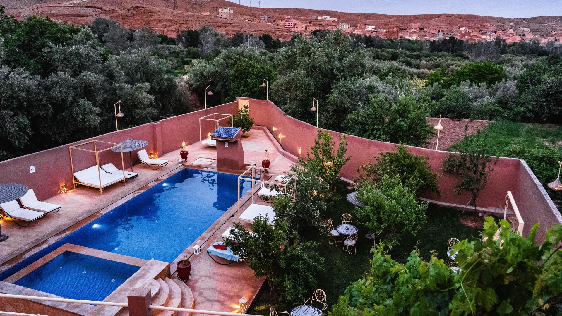Vista desde un ángulo elevado de una piscina rectangular azul rodeada por una pared de color terracota, tumbonas y frondosos árboles.
