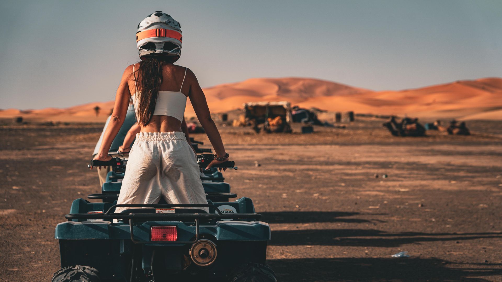Una persona con casco está sentada en un vehículo todoterreno en un paisaje desértico con grandes dunas de arena al fondo.